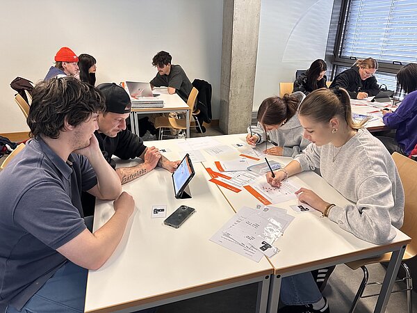 Mehrere Studierende sitzen in Gruppen an Tischen, bearbeiten Arbeitsblätter und nutzen digitale Geräte für eine gemeinsame Aufgabe.
