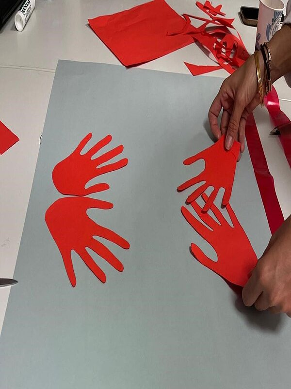 Eine Person bastelt mit roten Papp-Handabdrücken auf einem hellblauen Hintergrund. Links liegen zwei bereits ausgeschnittene Handabdrücke, während rechts eine Hand gerade platziert wird.