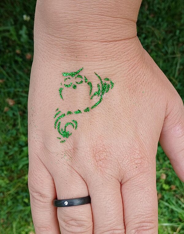 Ein grünes Glitzer-Tattoo in Form eines abstrakten Musters auf dem Handrücken einer Person.