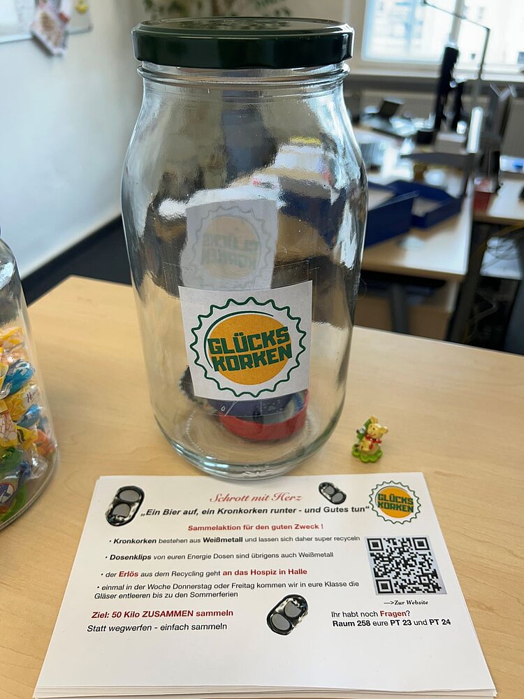 Sammelaktion: Kronkorken für den guten Zweck Ein großes, leeres Einmachglas mit grünem Deckel steht auf einem Tisch. Auf dem Glas klebt ein „Glückskorken“-Logo. Davor liegt ein Flyer mit Informationen zur Aktion „Schrott mit Herz“. Im Hintergrund ist ein Büro mit einem Schreibtisch und Fenstern zu sehen.
