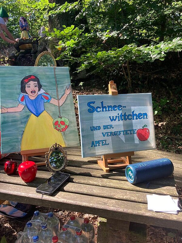Zwei kleine Leinwandbilder sind auf einen Holztisch im Wald aufgestellt. Auf einem ist Schneewittchen aus der Disney-Zeichentrickfilm zu sehen. Auf der anderen Leinwand steht "Schneewittchen und der vergiftete Apfel" geschrieben und es ist ein roter Apfel darauf gemalt. 
