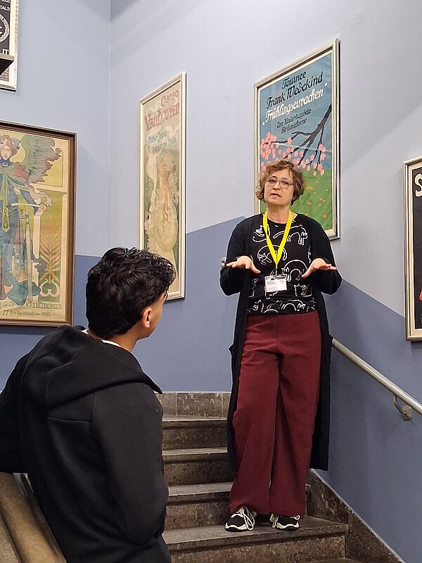 Museumsführerin mit gelbem Ausweisband spricht auf einem Treppenabsatz mit blauen Wänden zu einem Besucher; um sie herum hängen verschiedene historische Plakate in Rahmen.