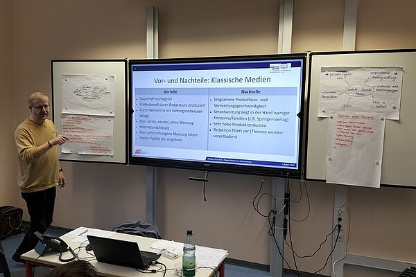 Seminarraum mit moderner Präsentationstechnik: Im Vordergrund steht eine männliche Person, die auf eine große digitale Präsentationsfläche zeigt. Die Präsentation trägt den Titel 'Vor- und Nachteile: Klassische Medien' und listet in zwei Spalten die jeweiligen Vorteile und Nachteile auf. Links sind Vorteile wie 'dauerhaft verfügbar', 'hohe Qualität' und 'breite Zielgruppe' aufgeführt, rechts Nachteile wie 'lange Produktionszeiten' und 'hohe Kosten'. Die Präsentation ist Teil des Projekts 'Werde zum Faktenchecker', was durch das Logo oben rechts und die Kontextangabe bestätigt wird. Links und rechts der Präsentationsfläche hängen große Papierbögen mit handschriftlichen Notizen und Diagrammen, die zusätzliche Informationen zum Thema bieten. Auf dem Tisch vor der Präsentationsfläche steht eine Wasserflasche. Die Szene vermittelt eine interaktive Lernatmosphäre mit Fokus auf Medienkompetenz und Faktenprüfung.