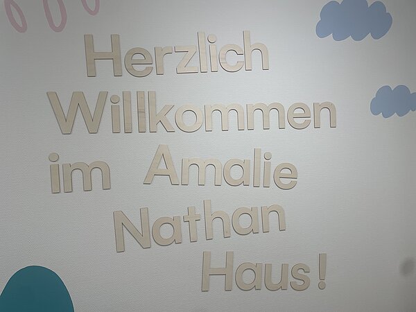 Auf einer hellen Wand mit aufgemalten Wolken steht in Holzbuchstaben der Schriftzug "Herzlich Willkommen im Amalie Nathan Haus!"