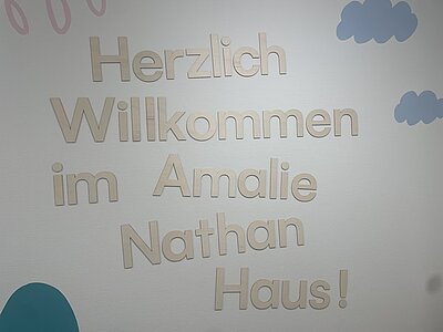 Auf einer hellen Wand mit aufgemalten Wolken steht in Holzbuchstaben der Schriftzug "Herzlich Willkommen im Amalie Nathan Haus!"