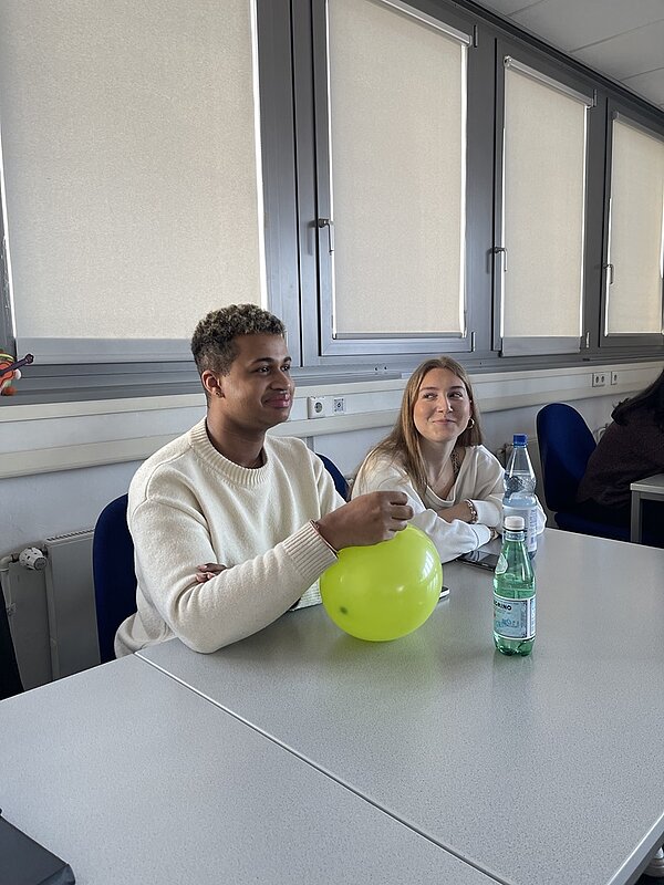 Zwei Auszubildende sitzen an einem Tisch in einem Zimmer mit großen, verdunkelten Fenstern. Eine Person hält einen leuchtend gelben Luftballon, die andere lächelt. Auf dem Tisch steht eine Flasche Mineralwasser. Beide tragen helle Pullover und scheinen in ein Gespräch oder eine andere Tätigkeit vertieft zu sein.