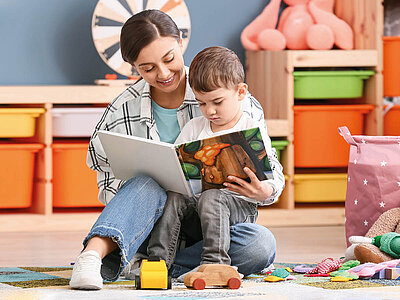 Eine Frau und ein Kind sitzen auf dem Boden in einem Raum, der wie ein Kinderzimmer oder ein Spielzimmer aussieht. Die Frau trägt ein kariertes Hemd und Jeans, während das Kind ein weißes T-Shirt und graue Hosen trägt. Sie lesen gemeinsam ein Bilderbuch, das das Kind auf dem Schoß hält. Um sie herum liegen verschiedene Spielzeuge, darunter ein gelbes Spielzeugauto und ein hölzernes Spielzeugauto. Im Hintergrund sind bunte Aufbewahrungsboxen und Regale mit weiteren Spielsachen zu sehen.
