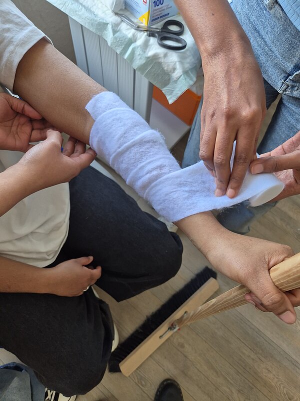 Teilnehmende legen gemeinsam einen Verband am Unterarm an und trainieren pflegerische Handgriffe.