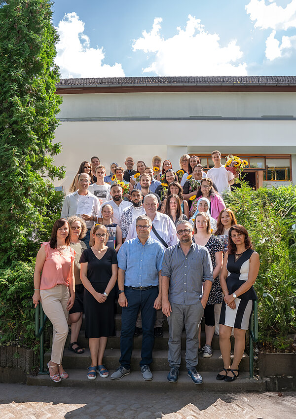 Feierliche Zeugnisvergabe der Pflegefachkräfte am 29. August 2024 Die Abschlussklasse Pflege 22 in Jena kommt für ein Gruppenbild zusammen. Eine Mischung von jungen Männern und Frauen posiert zusammen auf einer Treppe zwischen zwei Büschen.