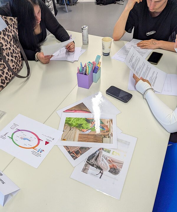 An einem Tisch studieren drei Schüler*innen verschiedene Infoblätter. Weiterhin befinden sich auf dem Tisch zwei Handys, ein Kaffeebecher, ein Energydrink sowie in der Mitte zwei Boxen mit Stiften.