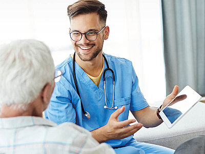 Ein Krankenpflegehelfer zeigt einem älteren Patienten auf einem Tablet etwas, während sie miteinander sprechen. Eine moderne Herangehensweise an die Patientenversorgung. 