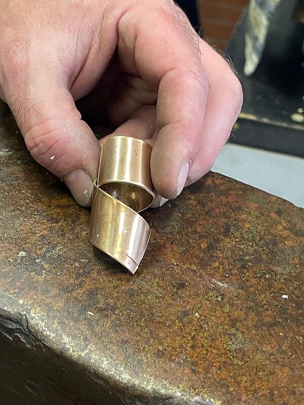 Nahaufnahme einer Hand, die ein gedrehtes goldfarbenes Metallstück über einer kupferfarbenen Oberfläche, wahrscheinlich die eines Ambosses, hält.