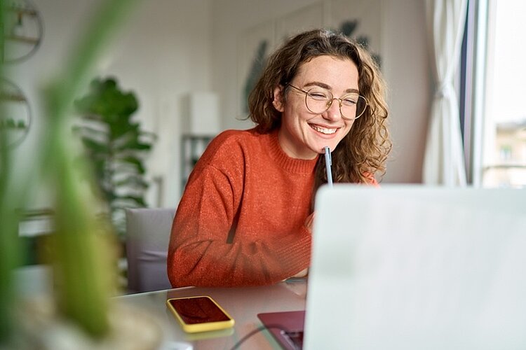 Lächelnde junge Frau sitzt vor dem Laptop am Schreibtisch, hält einen Stift in der Hand, während sie sich von zu Hause aus Webinare ansieht, online lernt, an virtuellen Veranstaltungen, Kursen oder Telefonkonferenzen teilnimmt.