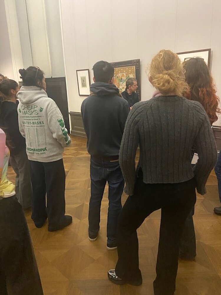 Eine Gruppe von Auszubildenden der Fremdkorrespondenten-Klasse der Euro Akademie Berlin steht dicht beieinander in einem historischen Ausstellungsraum der Alten Nationalgalerie in Berlin. Sie sind von hinten zu sehen und hören aufmerksam einer Museumsführung zu, die vor gerahmten Gemälden an der Wand stattfindet. Der Raum ist geprägt von klassischen Wandvertäfelungen, Parkettboden und traditioneller Hängung der Kunstwerke. Die Exkursion dient der kultur- und medienpädagogischen Auseinandersetzung mit bildender Kunst und journalistischer Beobachtung. Die Szene vermittelt eine konzentrierte Lernatmosphäre und zeigt praxisnahes Lernen im Museumskontext.
