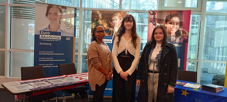 Drei Frauen stehen lächelnd vor einem Messestand, im Hintergrund sind Roll-Ups der Euro Akademie Bamberg zu sehen.