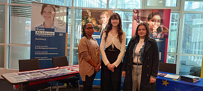 Drei Frauen stehen lächelnd vor einem Messestand, im Hintergrund sind Roll-Ups der Euro Akademie Bamberg zu sehen.