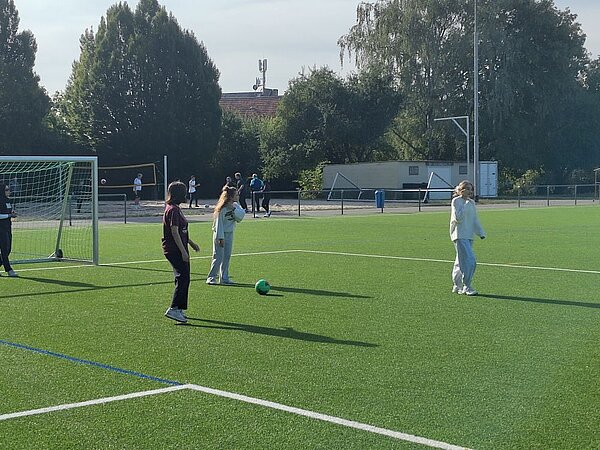 Eine Gruppe von Menschen steht auf einem grünen Kunstrasenplatz und spielt Fußball. Im Hintergrund sind Bäume, Gebäude und ein Fußballtor zu erkennen.