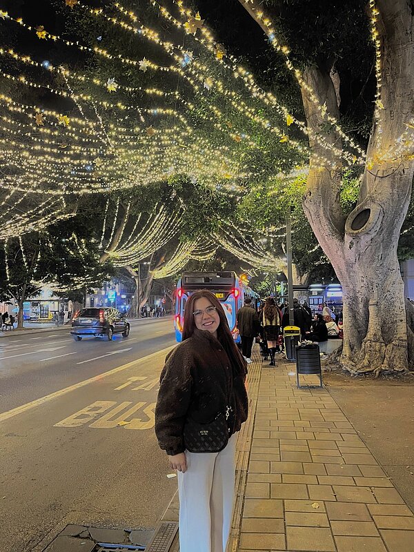 Junge Frau steht abends lächelnd an einer Bushaltestelle in Málaga, über der Straße spannt sich ein dichtes Dach aus warmweißen Lichterketten und Sternen.
