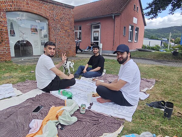 Drei Männer sitzen auf Picknickdecken im Grünen neben dem Schulgebäude, ein kleiner Hund steht daneben.