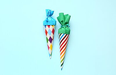 Zwei Schultüten liegen auf eine türkisfarbenen Fläche. Die linke ist etwas kleiner, hat ein Motiv mit bunten Rauten und einen hellblauen Deckel. Die rechte Schultüte ist bunt-gestreift mit einem grünen Deckel.