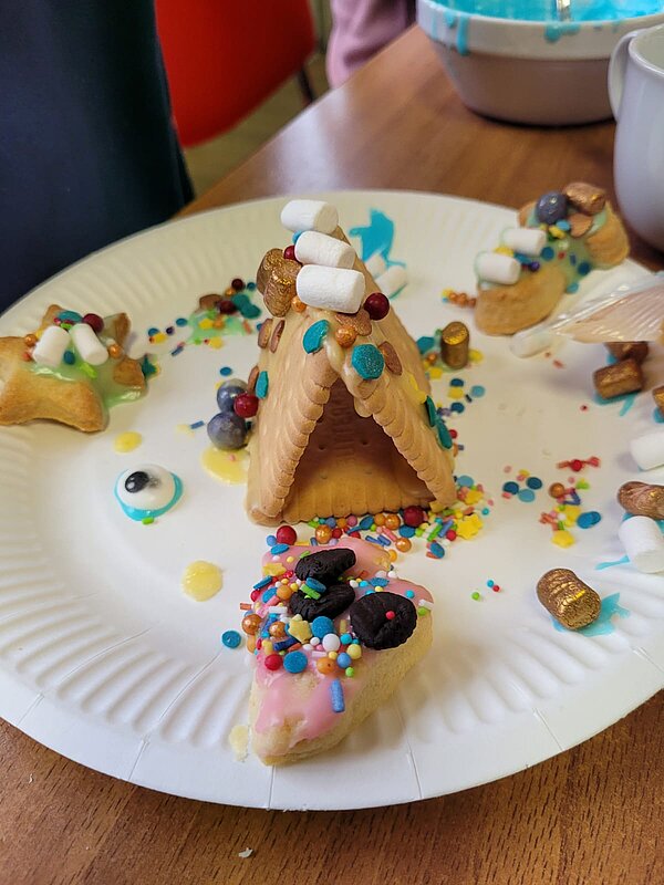 Ein selbstgemachtes Mini-Häuschen aus Butterkeksen auf einem Pappteller, dekoriert mit Zuckerguss, Mini-Marshmallows und bunten Streuseln.