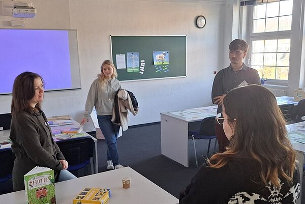 Tag der offenen Tür an der Euro Akademie Hohenstein-Ernstthal: Ein Klassenzimmer mit vier Personen, die sich unterhalten. Eine Person hält eine Jacke im Arm. Im Hintergrund sind ein Whiteboard, ein Fenster und eine grüne Magnettafel, an der verschiedene Dokumente hängen, zu sehen.