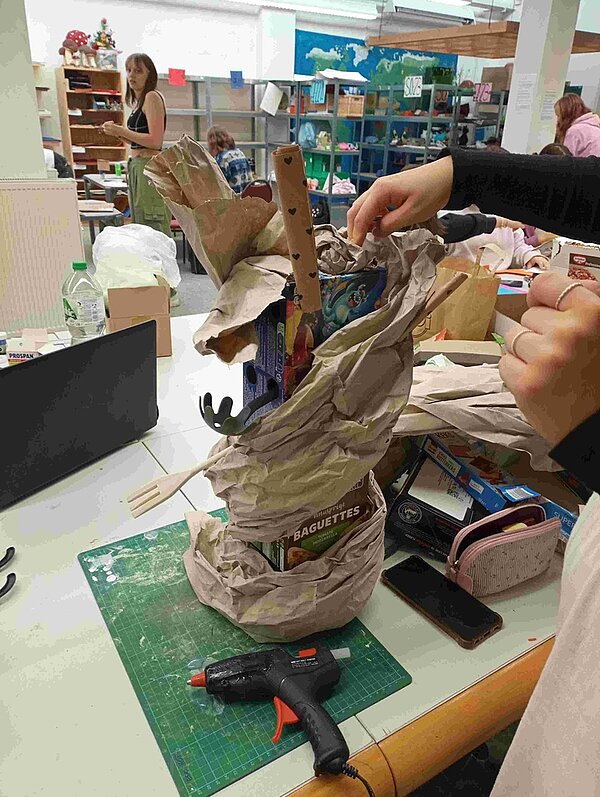 Blick in den Werkraum, der einen Tisch mit Bastelmaterialien und zwei Hände zeigt, die aus Abfall etwas Neues gestalten.