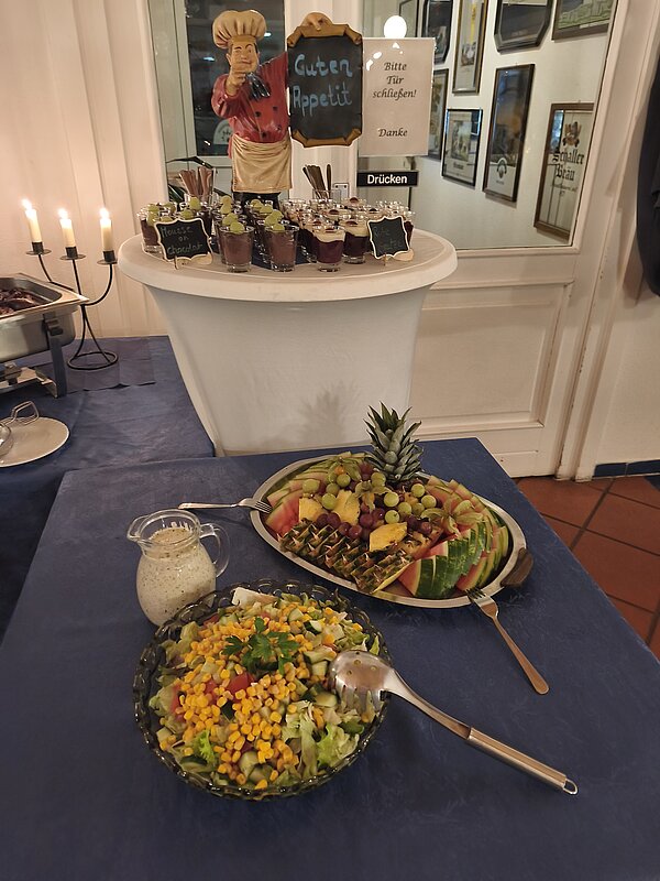 Buffet mit „Guten Appetit“-Schild: Dessertgläser, Kerzenleuchter, großer Obstteller und Schüssel grüner Salat mit Dressing.