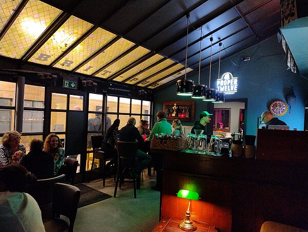 Blick in einen gemütlich beleuchteten Pub: Gäste sitzen an Tischen und an der Bar, über der Theke hängen Lampen, im Hintergrund leuchtet ein „Proper Twelve“-Neonschild.