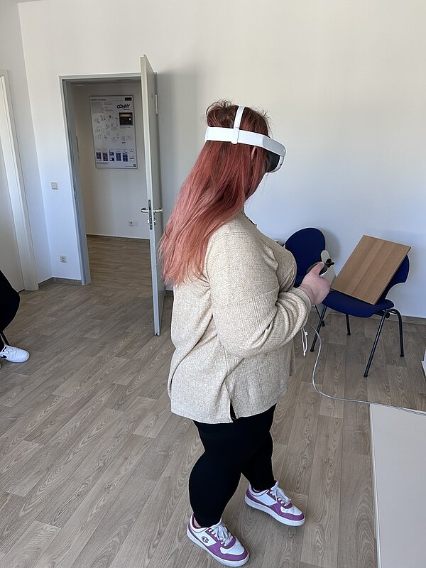 Eine Auszubildende mit langen, pinken Haaren trägt eine VR-Brille und benutzt einen Controller in einem Raum mit hellem Holzboden. Sie ist mit einem beigen Pullover, schwarzen Leggings und pink-weißen Turnschuhen bekleidet. Der Raum hat offene Türen, an der Wand hängt ein Poster, und im Hintergrund sind Stühle und ein Tisch zu sehen.