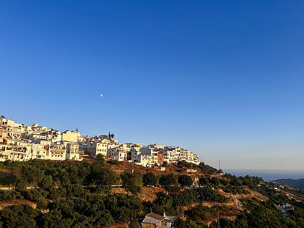 Erasmus-Bericht von Vanessa (Córdoba, 08/2024) Eine malerische Aussicht auf ein traditionelles weißes Dorf, das sich an einen Hügel schmiegt. Die weiß getünchten Häuser leuchten in der Abendsonne, während das tiefblaue Meer in der Ferne sichtbar ist. Im Vordergrund sind grüne Bäume und Felder zu sehen.