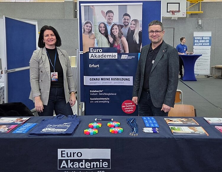 Zwei Mitarbeitende der Euro Akademie Erfurt stehen hinter einem Informationsstand auf der Berufsinformationsmesse am Staatlichen Berufsschulzentrum Arnstadt-Ilmenau. Vor ihnen befindet sich ein Tisch mit Informationsmaterialien wie Flyern, Broschüren, Schlüsselbändern, Give-aways und Stofftaschen mit dem Logo der Euro Akademie. Im Hintergrund ist ein großer Roll-up-Banner zu sehen, der Ausbildungsangebote wie Erzieher/in und Sozialassistent/in sowie praxisnahe und persönliche Bildungswege bewirbt. Die Messe findet in einer Sporthalle statt, erkennbar an Basketballkorb und Hallenboden. Die Szene zeigt eine freundliche Beratungssituation und vermittelt Einblicke in Berufsorientierung, Ausbildungsmöglichkeiten und Bildungsberatung.
