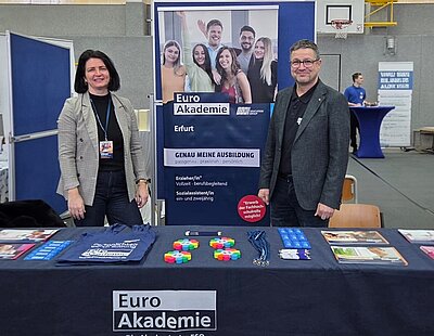 Zwei Mitarbeitende der Euro Akademie Erfurt stehen hinter einem Informationsstand auf der Berufsinformationsmesse am Staatlichen Berufsschulzentrum Arnstadt-Ilmenau. Vor ihnen befindet sich ein Tisch mit Informationsmaterialien wie Flyern, Broschüren, Schlüsselbändern, Give-aways und Stofftaschen mit dem Logo der Euro Akademie. Im Hintergrund ist ein großer Roll-up-Banner zu sehen, der Ausbildungsangebote wie Erzieher/in und Sozialassistent/in sowie praxisnahe und persönliche Bildungswege bewirbt. Die Messe findet in einer Sporthalle statt, erkennbar an Basketballkorb und Hallenboden. Die Szene zeigt eine freundliche Beratungssituation und vermittelt Einblicke in Berufsorientierung, Ausbildungsmöglichkeiten und Bildungsberatung.