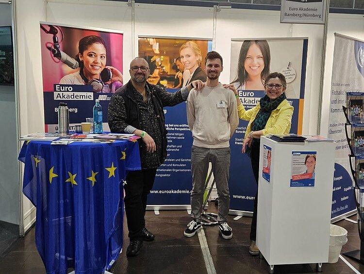 Drei gut gelaunte Personen stehen am Stand der Euro Akademie Bamberg-Nürnberg auf der Studienmesse Stuzubi 2025. Im Hintergrund sind Informationsbanner mit lächelnden Studierenden und dem Logo der Akademie zu sehen. Ein Stehtisch mit einer EU-Flagge sowie ein Infotresen mit Flyern vervollständigen das Messe-Setup.