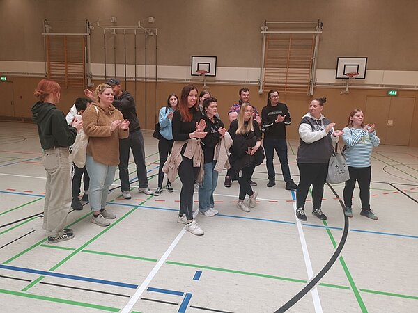 Kita-Sportfest Eine Gruppe von etwa fünfzehn Personen steht in einer Turnhalle. Die Personen, hauptsächlich junge Erwachsene, stehen in zwei kleinen Gruppen und scheinen zu klatschen. Im Hintergrund sind Basketballkörbe und Turngeräte an der Wand zu sehen. Die Halle hat Markierungen auf dem Boden für verschiedene Sportarten.