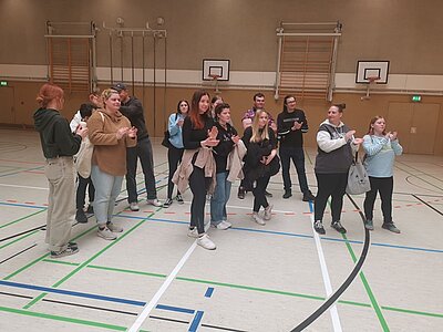 Eine Gruppe von etwa fünfzehn Personen steht in einer Turnhalle. Die Personen, hauptsächlich junge Erwachsene, stehen in zwei kleinen Gruppen und scheinen zu klatschen. Im Hintergrund sind Basketballkörbe und Turngeräte an der Wand zu sehen. Die Halle hat Markierungen auf dem Boden für verschiedene Sportarten.