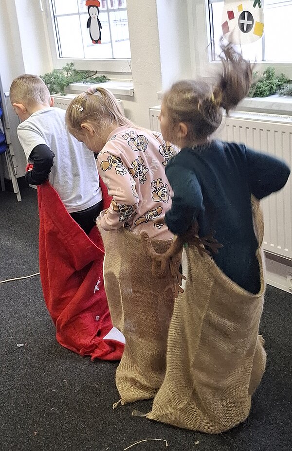 Mehrere Kinder hüpfen in Jutesäcken durch einen Raum, in dem weihnachtliche Fensterdekorationen hängen.
