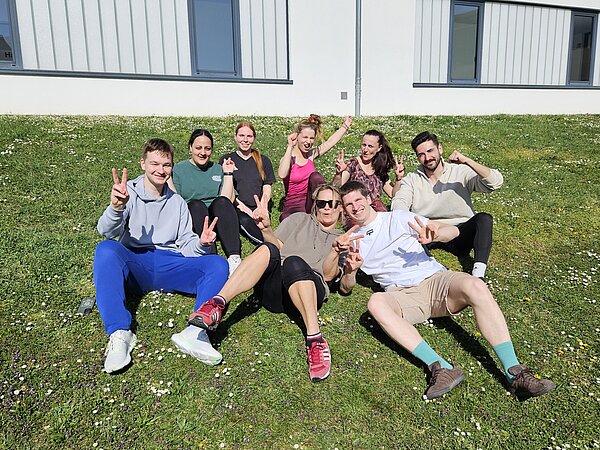 Eine Gruppe junger Menschen in Sportkleidung sitzt gutgelaunt auf einer Wiese vor einem modernen Gebäude. Sie lachen, zeigen Friedenszeichen und wirken entspannt und zufrieden.