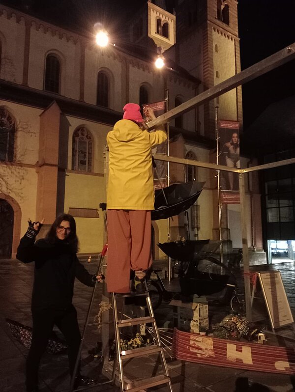 Im Dunkeln hängen zwei Menschen vor dem Dom an einem Rahmen einen Regenschirm auf.