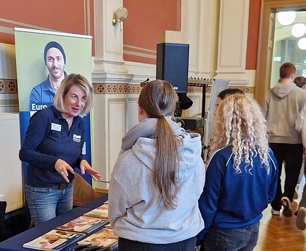 Mitarbeiterin am Messestand der Euro Akademie in angeregtem Beratungsgespräch mit zwei interessierten Schülerinnen.