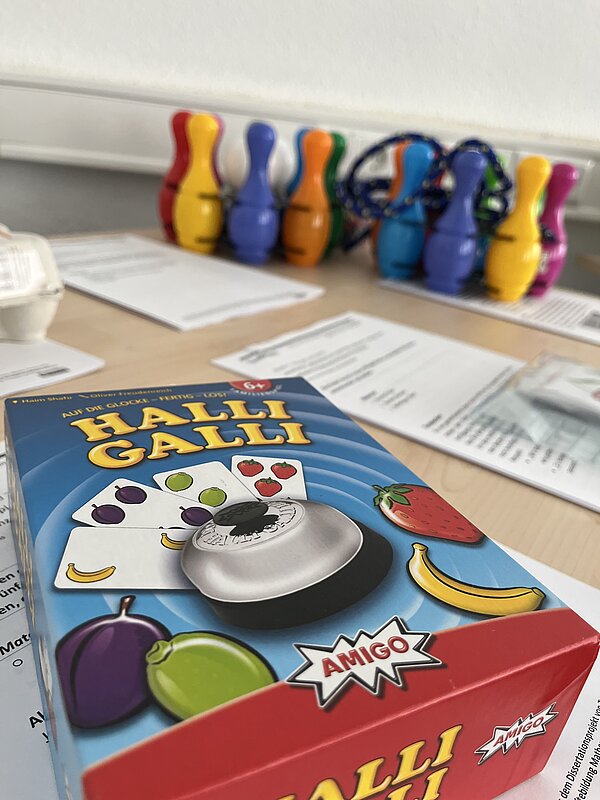 Verpackung des Spiels „Halli Galli“ mit farbenfrohen Obstsymbolen und Glocke auf einem Tisch. Im Hintergrund stehen bunte Mini-Kegel und weitere Lernmaterialien.