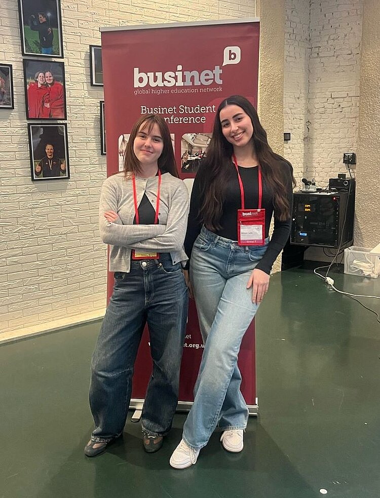Zwei junge Frauen stehen lächelnd vor einem Businet-Banner mit der Aufschrift „Businet Student Conference“. Sie tragen rote Namensschilder und bequeme Jeans-Outfits, posieren mit verschränkten Armen und wirken selbstbewusst und glücklich. Im Hintergrund sind Bilder und eine Backsteinwand zu sehen.
