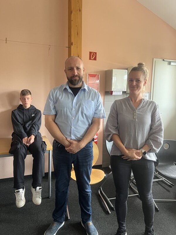 Gutgelaunter gemeinsamer Start ins Schuljahr Zwei Lehrkräfte stehen in einem Raum und stellen sich scheinbar vor. Ein Schüler sitzt im Hintergrund auf einem Tisch.