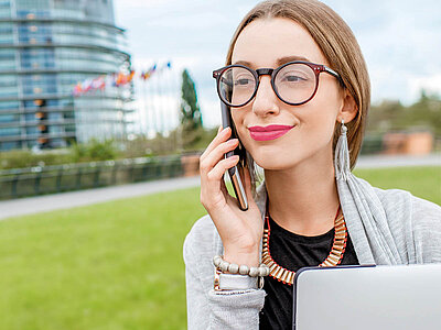 Eine Frau mit Laptop und Handy steht vor einem modernen Gebäude mit Flaggen.