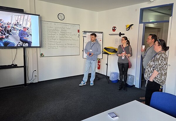 Erfolgreicher Tag der offenen Tür Besucher*innen stehen in einem Klassenraum, haben etwas zu trinken in der Hand und schauen sich auf einem großen Bildschirm eine Diashow an.