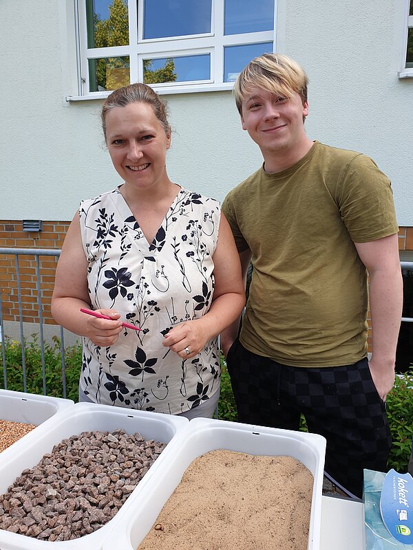 Ein erlebnisreicher Vormittag voller Spiel, Kreativität und Gemeinschaft Eine Frau und ein junger Mann stehen an einem Tisch mit verschiedenen Materialien wie Sand, Steinen und Erde für eine Fühlstation.