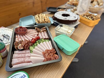 Ein Büfett mit einer Auswahl an Speisen. Zu sehen sind verschiedene Wurstsorten, Gurken, Tomaten und Feigen. Daneben stehen kleine Behälter mit Dips und weitere Snacks wie Knabbereien und ein Kuchen.