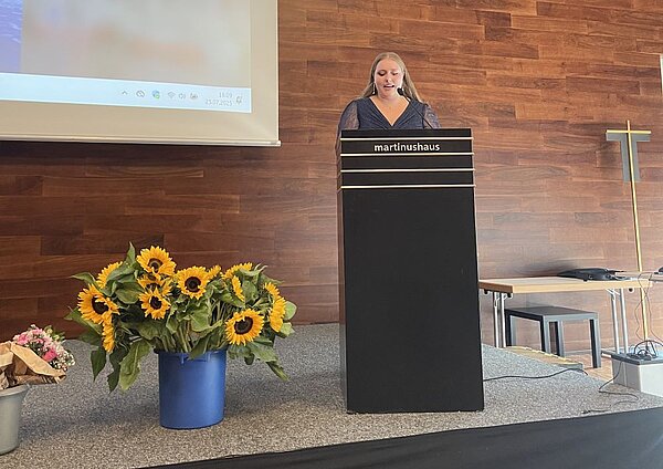 Eine Frau steht an einem Pult mit der Aufschrift "martinushaus" und hält eine Rede. Auf dem Podium stehen Blumenarrangements mit Sonnenblumen. Im Hintergrund ist eine Leinwand mit einer Präsentation zu sehen.