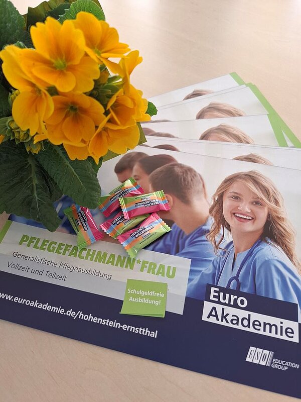 Mehrere Flyer der Euro Akademie liegen auf einem Tisch. Daneben steht eine Vase mit gelben Blumen. Auf den Flyern ist eine Frau im Pflegeberuf abgebildet.