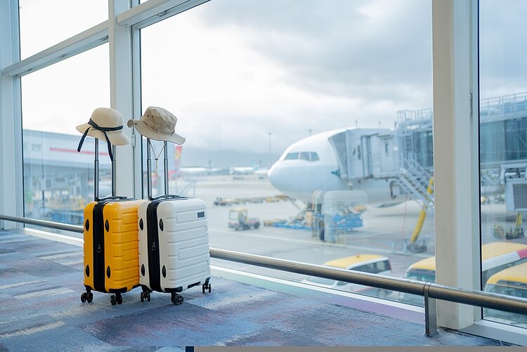 Berufspraktikantin Alexandra Verl berichtet von ihrem Auslandsaufenthalt in Malaga Zwei Koffer mit Sonnenschutzhüten stehen in einem Flughafen vor einem Fenster. Dahinter sieht man ein parkendes Flugzeug.